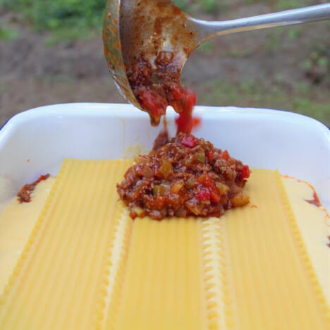 Making lasagna with a béchamel sauce topped with sheets of lasanga noodles and a ladle of Bolognese sauce being added.