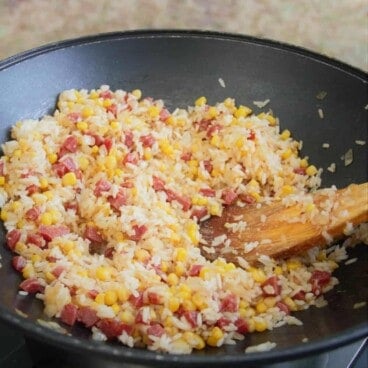 A wooden spoon stir fries a mixture of rice, corn, and Chinese sausage in a large wok.