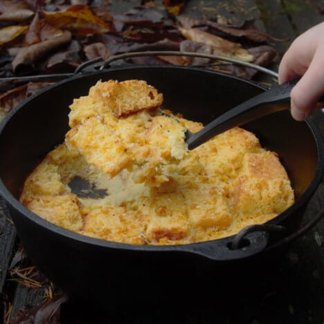 A hand holding a serving spoon full of baked, golden brown corn pudding over a Dutch oven containing the rest of the pudding.