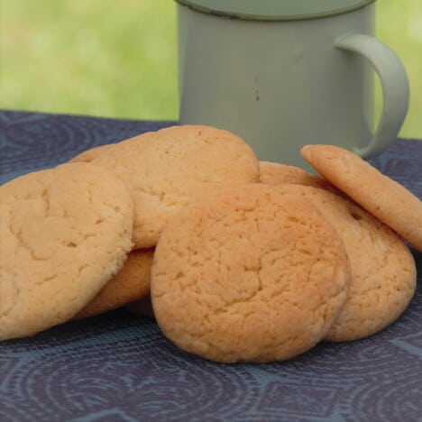 A pile of golden brown cream cheese cookies rests on a blue napkin next to a green mug.