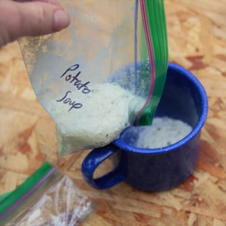 A zip top bag of powdered potato soup mix pouring into a blue camping mug.