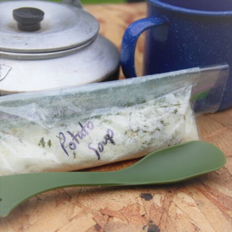 A homemade zip top bag of potato soup sitting on a board with a camp mug, spoon, and kettle.
