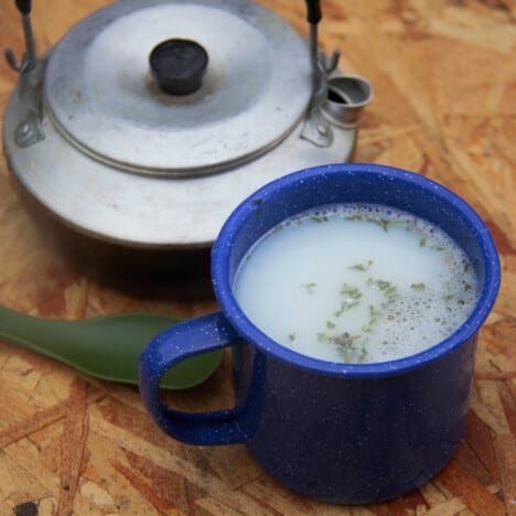 A blue camp enamel mug filled with the potato soup, behind it is a Trangia light-weight camp kettle.