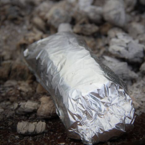 A pizza roll completely wrapped in foil sits on a bed of hot coals.