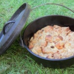 A Dutch oven sitting on the grass with a cooked hot cross bun pudding inside.