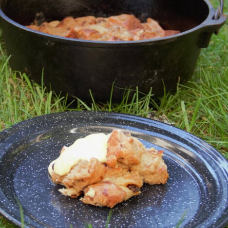 A black camp plate with a serving of hot cross bun pudding and custard with the Dutch oven and remaining pudding in the background.