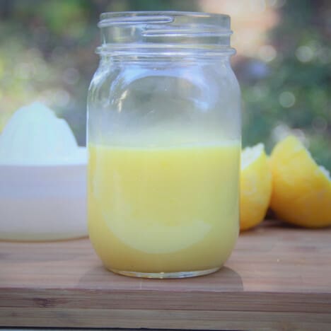 A half jar of lemon curd sitting on a wooden chopping board with squeezed half lemons in the background.