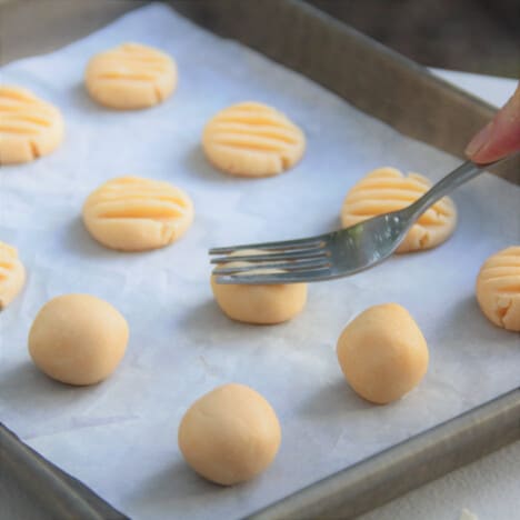 A baking paper-lined baking tray with balls of dough being flattened with a fork.