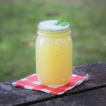 A pineapple mint mojito mocktail served in a jar garnished with mint, sitting on a red and white checkered napkin on a picnic table.