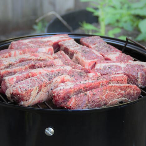 Raw short ribs are on the grill grate of a smoker.