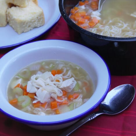 A camp bowl of turkey noodle soup next to the black pot containing more soup.