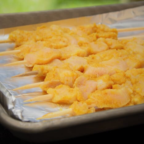 Raw chicken satay are lined up on a foil-lined tray ready to be cooked.