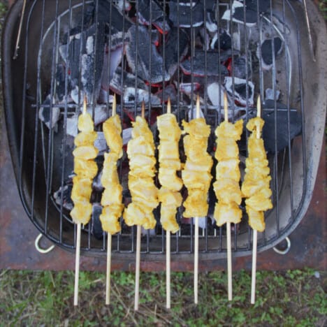 Looking down on a small grill with the satay skewers on it with the chicken over the heat and the exposed skewers hanging out of the grill so they don't burn.