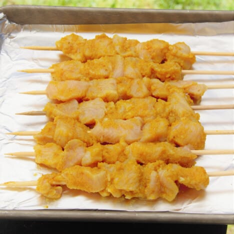 Looking down onto a foil-lined baking tray filled with uncooked chicken satay skewers.