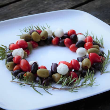 Looking into a antipasto Christmas wreath on a white platter, with mozzarella, olives, tomatoes, and rosemary sprigs.