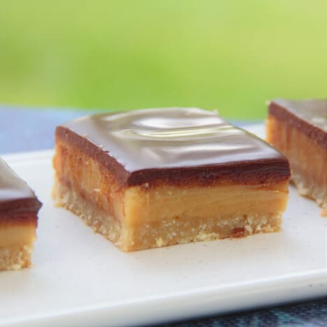 A close up of a square of caramel slice on a serving platter, with distinct layers of cookies, caramel, and chocolate.