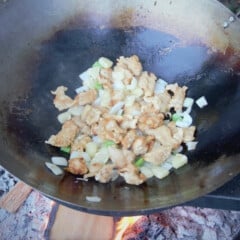 A wok is over a campfire with the chicken, pineapple, onion, and bell pepper stir frying.