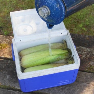 A small blue six-pack cooler with four ears of corn in it being filled with boiling water.