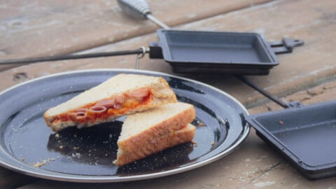 Looking down onto an untoasted ham and pineapple sandwich, cut in half, on a black camping plate on a picnic table with the pie iron next to the plate.