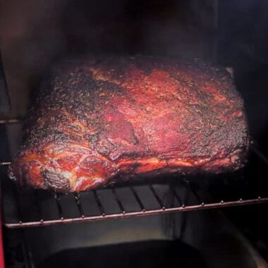 A shoulder of lamb in a smoker part way through the cook with a mahogany tone.