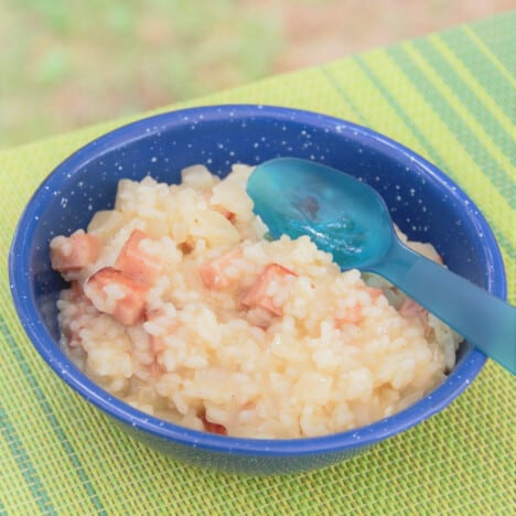 A blue camp bowl of spam risotto sitting with a blue camp spoon ready to be eaten.