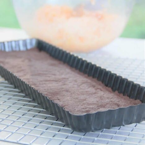 A baking tin on a cooling rack lined with the cookie base, with a glass bowl of the filling in the background.