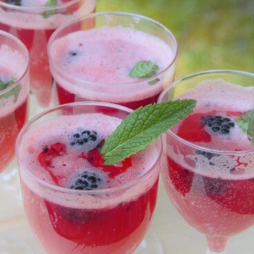 A batch of blackberry summer mocktails are lined up on a table, the main one garnished with two blackberries and a mint leaf.