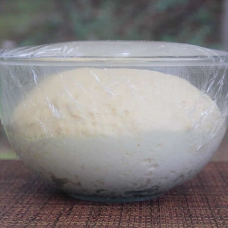 A clear glass bowl covered with plastic wrap and a fully risen dough inside it.
