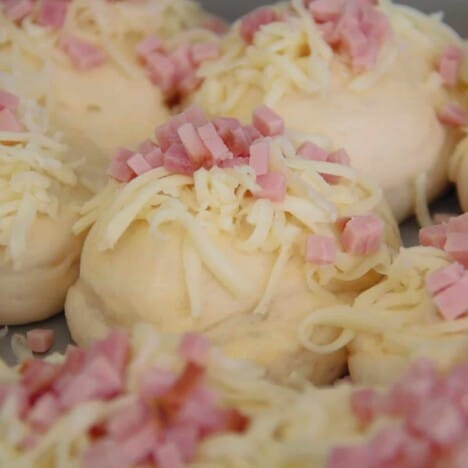 A close up shot of a proofed but not baked cheese and bacon roll with shredded cheese and diced ham on top.