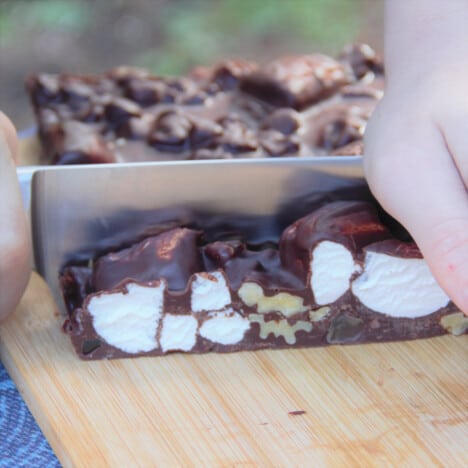 The slice is being cut into squares on a wooden chopping board, with large chunks of nuts and marshmallows visible.