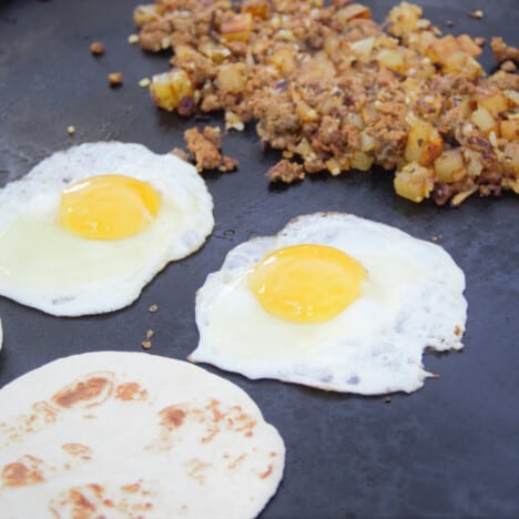 Looking accross a flat top grill showing the tortilla, fried eggs and the chorizo, potato, and onion hash.