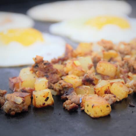 A flat top grill showing the chorizo and potato hash with the fried eggs and tortillas in the background.