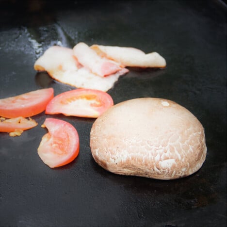 A flat top grill with a large portobello mushroom upside-down, tomatoes, and bacon.