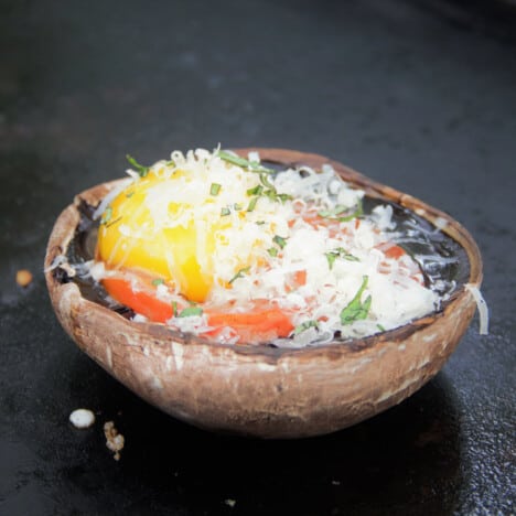 A portobello mushroom upside down on a flat top grill filled with tomato and egg, then topped with cheese and parsley waiting to be cooked.