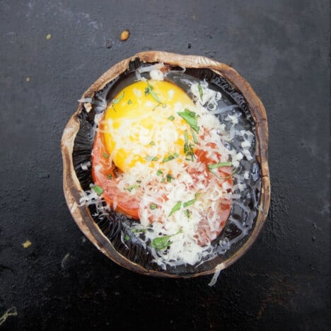 Looking down on a flat top grill with a mushroom recently filled with an egg then sprinkled with cheese and parsley.