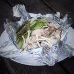 An open foil pack is sitting on a white plate showing its cooked contents of chicken, mushrooms, and bok choy.