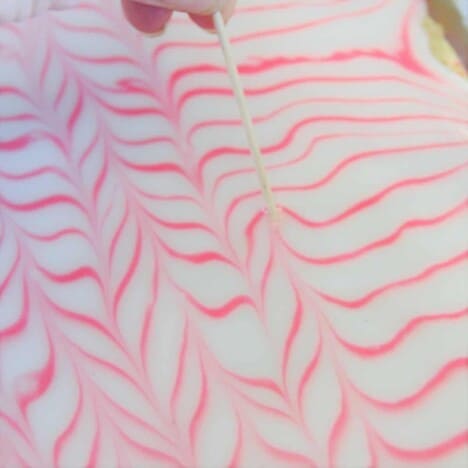 A toothpick is being dragged through the pink lines of icing on the vanilla slice to create a decorative pattern.