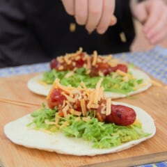 Shredded cheese being sprinkled over a hot dog taco on a wooden cutting board.