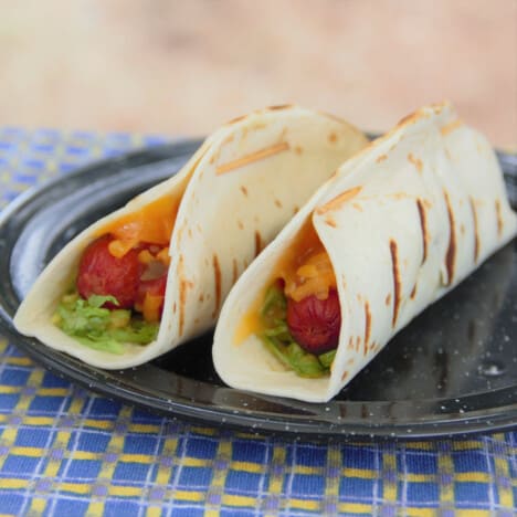 Two grilled hot dog tacos sit on a black camping plate on a blue and yellow checkered tablecloth.