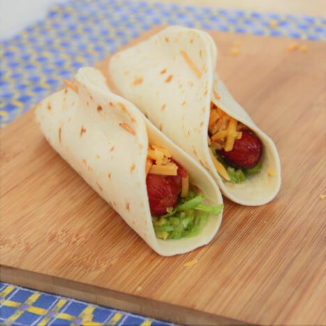 A chopping board with two hot dog tacos ready to cook.