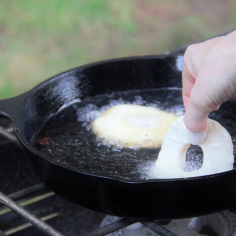 A skillet on a camp stove with a cooking pineapple fritter and a second one gently being added to the oil.