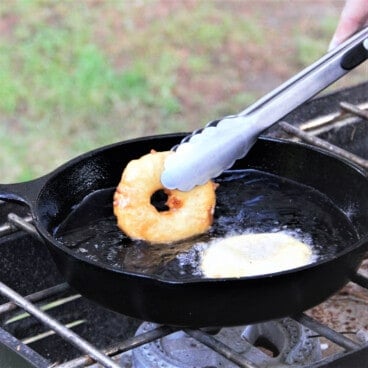 A skillet on a camp stove deep frying two pineapple fritters, one white and recently battered, the other golden brown and being turned with tongs.