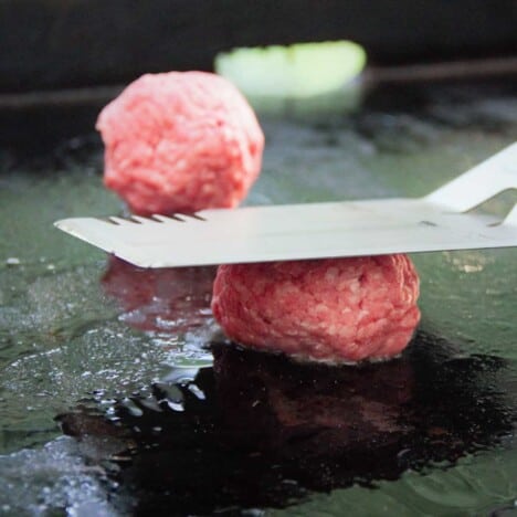 A flat top grill with two balls of ground beef, one being squashed flat with a large BBQ spatula.