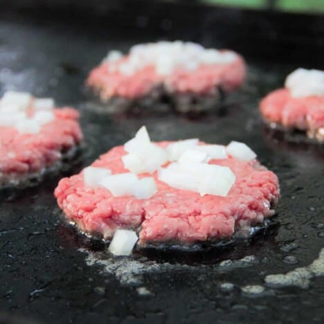 A flat top grill with recently added and pressed smash burgers with the onions already added to the top.