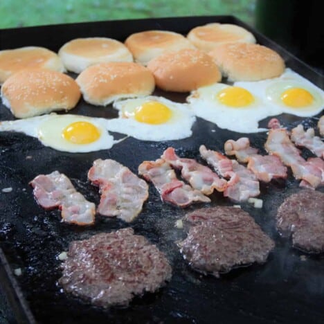 A fully loaded flat top grill with lines of smash burger patties, bacon, eggs, and hamburger buns.
