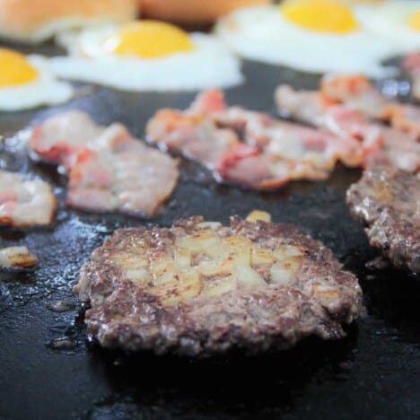 A flat top grill with the focus on a smash burger with bacon, eggs, and buns in the background.