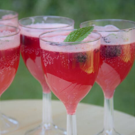 A row of blackberry summer mocktails in wine glasses, garnished with blackberries and mint, sit on a wooden table.