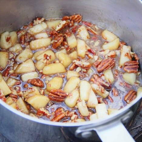 A stainless steel cooking pot is filled with chopped apples and pecans in a butter and maple syrup sauce.