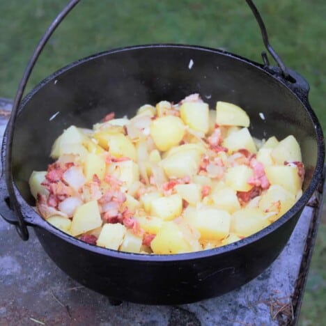 A Dutch oven sitting on concrete filled with bacon potatoes.