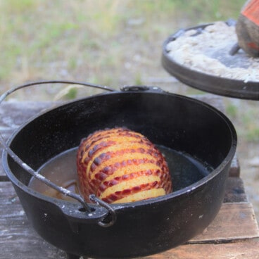 A Dutch oven with the charcoal laden lid being removed to expose the ham and pineapple within.